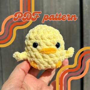 May include: A crocheted yellow chick amigurumi toy with blue eyes and an orange beak. The toy is being held in a hand. The background is a retro-style pattern of orange and brown stripes.