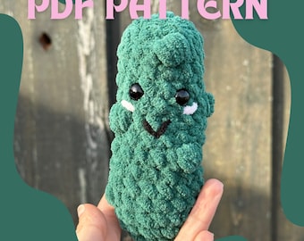 May include: A green, plush, crocheted pickle with black eyes, a black smile, and white accents. The text "PDF PATTERN" is displayed above the pickle. The pickle is held in a hand against a wooden background.