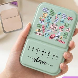 May include: A sage green rectangular compact mirror featuring a collection of religious affirmations and the text "MY DAILY BIBLE AFFIRMATIONS" on the top half, with a row of seven black crosses and the name "gloria" printed on the bottom half.