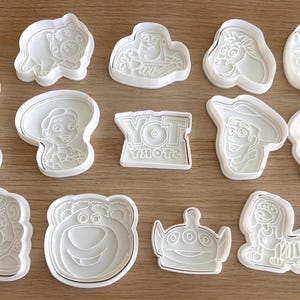 May include: A selection of white plastic cookie cutters depicting characters from the animated film "Toy Story." The cutters include Buzz Lightyear, Woody, Jessie, and others. The word "TOY STORY" is also featured in a cutter.