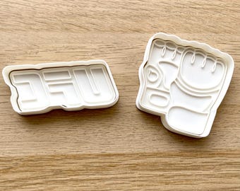May include: Two white plastic cookie cutters. One cutter spells out the letters "DFU" and the other is shaped like a clenched fist. The cutters are on a light brown wooden surface.
