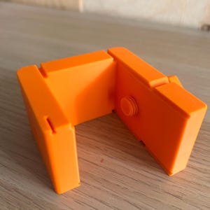 May include: A bright orange U-shaped plastic component featuring a smooth matte finish. The object consists of three connected rectangular segments with a small circular protrusion on the inner side of one panel and narrow vertical slots on the edges.
