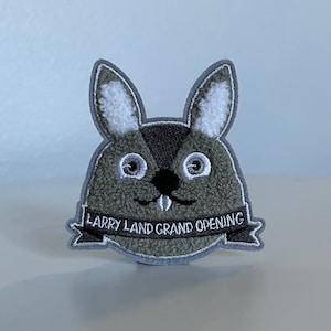 May include: A gray plush patch shaped like a rabbit with white fur ears and a black nose. The patch has the text "Larry Land Grand Opening" on a white banner.