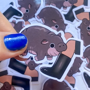 Puede incluir: Una pegatina con una ilustración de dibujos animados de un pequeño animal marrón parecido a un cerdo con la nariz rosa y ojos grandes. El animal está siendo pisado por una pierna humana que lleva una bota de lluvia negra.