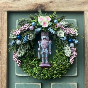 May include: A miniature Christmas wreath with a central nutcracker figurine. The wreath is made of green moss, decorated with pink and blue beads, white flowers, and glitter. The nutcracker is light blue and purple.