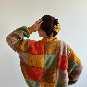 Peut inclure: Un pull en crochet à manches longues présentant un motif patchwork de carrés jaune moutarde, orange brûlé, vert sauge et beige. Le vêtement possède un col et des poignets en tricot côtelé, réalisés avec un point de crochet visible.