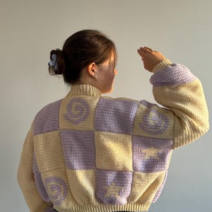 Puede incluir: Suéter de crochet con diseño de patchwork. El suéter tiene cuello alto y mangas largas. La combinación de colores incluye cuadrados de amarillo pálido y lavanda, con un diseño de carita sonriente en algunos cuadrados. El suéter se usa con vaqueros azules.