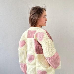 May include: A crocheted cardigan with a patchwork design featuring pink hearts on a cream background.