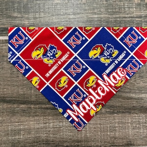 May include: A red and blue bandana with a repeating pattern of the University of Kansas logo and the letters "KU". The bandana is personalized with the name "Maple Mae" in white lettering.