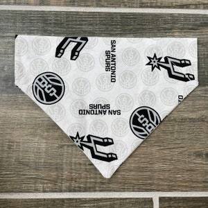 May include: White and grey San Antonio Spurs dog bandana. The bandana features the team's logo, the Spurs logo, and the words "San Antonio Spurs" in black. The bandana is made of a soft fabric and is triangular in shape.