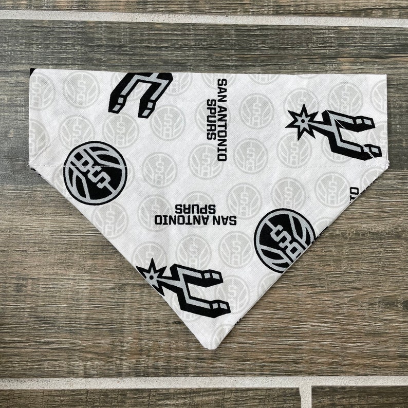 May include: White and grey San Antonio Spurs dog bandana. The bandana features the team's logo, the Spurs logo, and the words "San Antonio Spurs" in black. The bandana is made of a soft fabric and is triangular in shape.