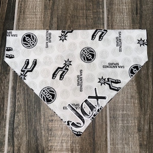 May include: White dog bandana with a repeating pattern of the San Antonio Spurs logo and text. The bandana has a personalized name, "Jax," in a bold, black font. The Spurs logo includes a basketball and a spur.