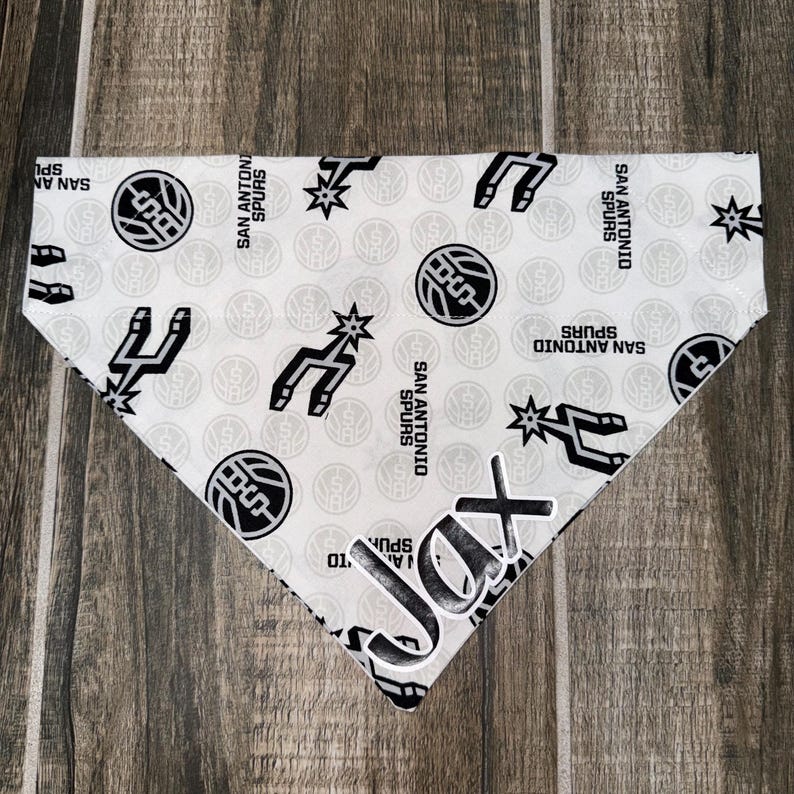 May include: White dog bandana with a repeating pattern of the San Antonio Spurs logo and text. The bandana has a personalized name, "Jax," in a bold, black font. The Spurs logo includes a basketball and a spur.