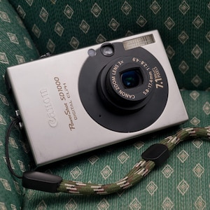 May include: Silver Canon PowerShot SD1000 digital camera with a black lens and a green and brown patterned wrist strap. The camera has a 3x zoom lens and 7.1 megapixels. The camera is resting on a green patterned surface.