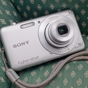May include: A silver Sony Cyber-shot digital camera featuring a 5x optical zoom lens, 16.1 megapixels, and a 3.2-6.5/5-25 lens specification. The compact device includes a built-in flash and a grey wrist strap attached to the side.