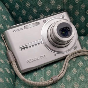 May include: A silver Casio Exilim digital camera featuring a prominent circular lens barrel on the right side, a built-in flash, and a textured beige wrist strap attached to the side. The brand name Casio and the model name Exilim are printed.