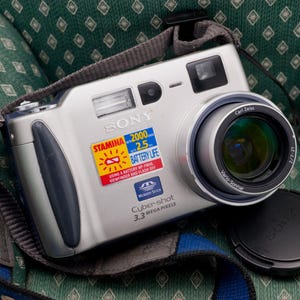 May include: Silver Sony Cyber-shot digital camera with a Carl Zeiss lens and a black lens cap. The camera has a strap and a sticker indicating 2.5 battery life and 3.3 megapixels. The camera is resting on a green patterned surface.