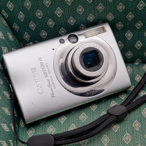 May include: A silver Canon PowerShot SD1100 IS Digital ELPH camera featuring a central lens, a built-in flash, and a black wrist strap. The camera body displays the brand name and model text clearly on the front metallic surface.