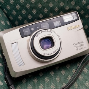 May include: A silver Nikon One-Touch Zoom 90s AF Quartz Date camera featuring a 38-90mm Macro zoom lens. The compact point-and-shoot device includes a built-in flash, a viewfinder, and a grey wrist strap attached to the side of the camera body.