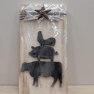 May include: A rustic wooden sign featuring a stack of farm animals: a cow, a pig, and a rooster. The sign is topped with a star and a blue and white striped bow. The sign is wrapped in clear plastic.