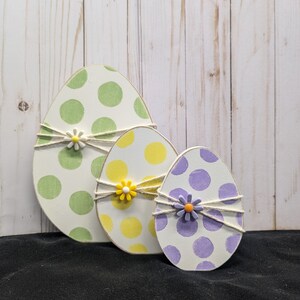 May include: Three wooden Easter eggs decorated with polka dots. The largest egg is green, the middle egg is yellow, and the smallest egg is purple. Each egg has a small white flower with a yellow center.