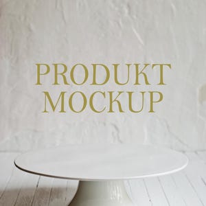 May include: A white ceramic cake stand with a wide, round top and a pedestal base. The stand is set against a textured white background and white wooden planks. The words "PRODUKT MOCKUP" are in gold lettering.