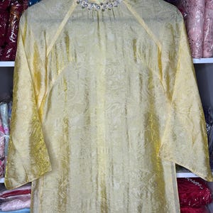 May include: A long, flowing yellow dress with a subtle paisley pattern. The dress features long, flared sleeves and a high neckline adorned with small, sparkling embellishments. The fabric has a slight sheen.