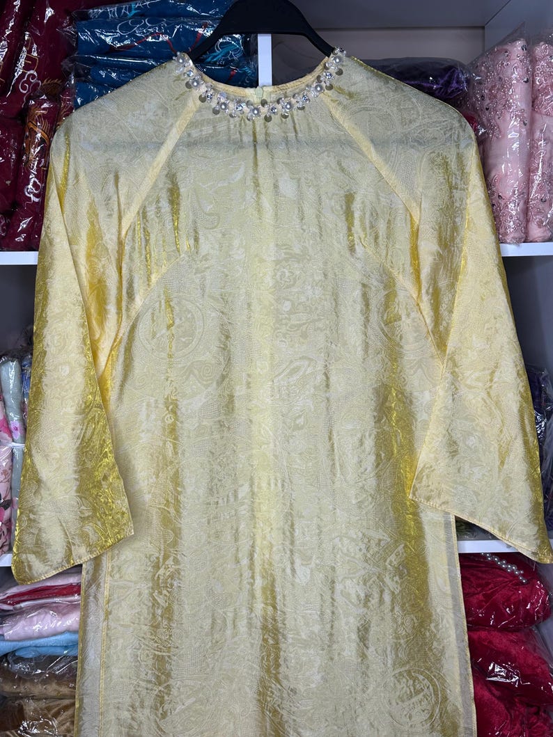 May include: A long, flowing yellow dress with a subtle paisley pattern. The dress features long, flared sleeves and a high neckline adorned with small, sparkling embellishments. The fabric has a slight sheen.