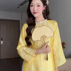 May include: A woman wearing a yellow traditional dress with long sleeves and a decorative collar. She is holding a beige fan. The dress has a subtle texture, and the overall look is elegant and cultural.