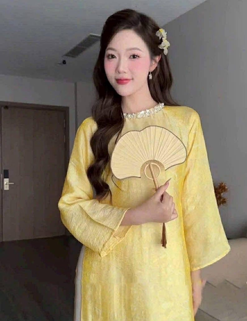 May include: A woman wearing a yellow traditional dress with long sleeves and a decorative collar. She is holding a beige fan. The dress has a subtle texture, and the overall look is elegant and cultural.