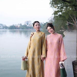 May include: Two women wearing traditional long dresses. One dress is yellow, the other is pink. Both dresses have long sleeves and mandarin collars. The women are standing near a body of water. The text "NGUYEN VAN TRINH" is visible.