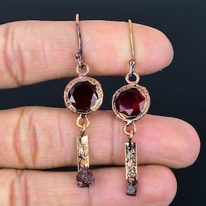 Op de afbeelding: Een paar koperen oorringen met een rode edelsteen en een kleinere rode edelsteen die eraf hangt. De oorringen zijn op een hand.