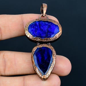 Könnte beinhalten: Eine Halskette mit Anhänger, bestehend aus zwei blauen Edelsteinen, die in Kupfer gefasst sind. Der größere Stein ist oval geformt, der kleinere tropfenförmig.