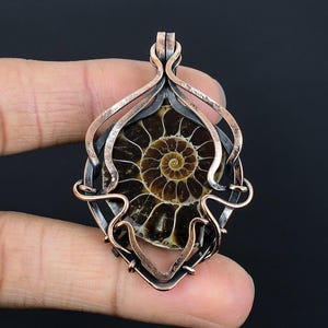 Pode incluir: Um pingente com um amonite fossilizado emoldurado em uma estrutura metálica decorativa. A amonite exibe um padrão espiral em tons de marrom e bege. A estrutura é uma combinação de metal prateado e cobre, criando um contraste marcante. O pingente é exibido em um fundo escuro.
