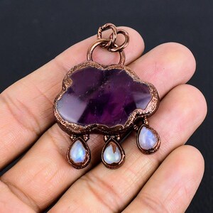 May include: A handcrafted pendant featuring a cloud-shaped amethyst stone with copper edging. Three teardrop-shaped moonstone charms dangle below. The pendant is held in a hand against a dark background.