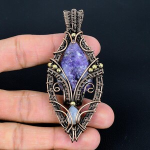 May include: A handcrafted pendant featuring a purple gemstone set within a copper wire frame. The wirework is intricately woven, with small gold-colored beads adding detail. The pendant has a teardrop shape, suitable for a necklace.