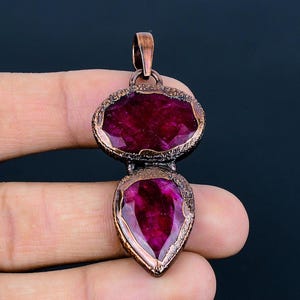 May include: A copper pendant with two ruby gemstones. The top gemstone is oval shaped and the bottom gemstone is teardrop shaped.