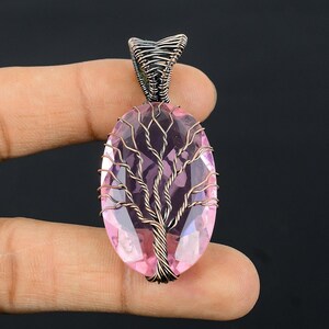 Puede incluir: Colgante artesanal con una piedra preciosa rosa de forma ovalada, con un dise&ntilde;o de &aacute;rbol envuelto en alambre. El colgante tiene una argolla plateada y lo sostiene una mano sobre un fondo oscuro. La piedra mide aproximadamente 3,8 cm de largo.