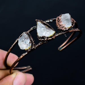 May include: A copper-colored cuff bracelet featuring three clear, irregularly shaped gemstones. The gemstones are set within a textured copper frame. The bracelet has an open design and is displayed against a dark background.