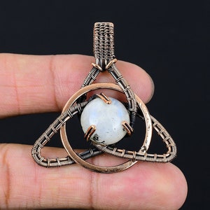 May include: A copper wire wrapped pendant featuring a round, white moonstone gemstone. The pendant is in the shape of a triangle with a circle around the stone.