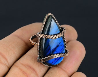 May include: A teardrop-shaped labradorite gemstone ring. The iridescent blue and green stone is set in a copper-colored, twisted wire frame. The ring has a simple band and a decorative setting that wraps around the stone.