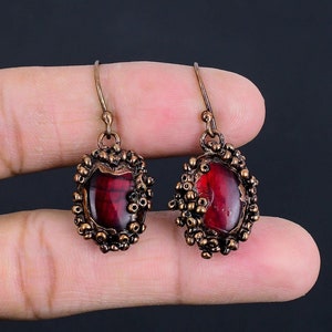 May include: A pair of dangle earrings featuring a red, oval-shaped gemstone set in a copper frame with a beaded design.