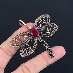 May include: A copper wire dragonfly pendant with a red gemstone in the center. The dragonfly is intricately detailed with wirework and has a loop at the top for hanging.