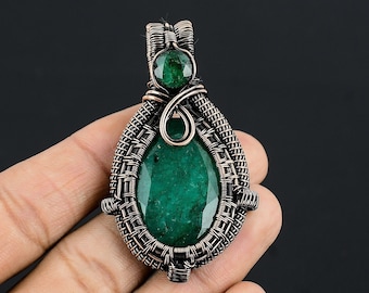 May include: A handcrafted pendant featuring a large, faceted emerald-green gemstone. The stone is encased in a woven copper wire setting, with a smaller emerald accent at the top. The pendant has a teardrop shape and is displayed against a dark background.