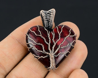 Puede incluir: Un colgante en forma de corazón con un diseño de árbol de la vida de alambre de cobre, engastado en una piedra preciosa roja. El colgante está hecho de alambre de cobre y una piedra preciosa roja.