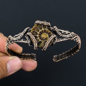 Könnte beinhalten: Ein Armband aus umwickeltem Kupferdraht mit einem versteinerten Ammoniten in der Mitte. Der Ammonit ist braun und hat ein spiralförmiges Muster.