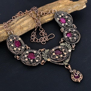 Puede incluir: Un collar de alambre de color cobre con cinco eslabones ovalados, cada uno con una piedra preciosa de color magenta intenso. El trabajo en metal muestra intrincados patrones de espirales y pequeños acentos circulares, unidos por una cadena con cierre de gancho.