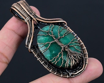 Puede incluir: Un colgante envuelto en alambre de cobre con una piedra preciosa verde con un diseño de árbol. El colgante tiene forma ovalada y tiene una anilla de cobre.