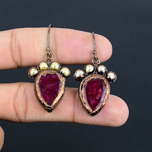 Può includere: Un paio di orecchini pendenti con una pietra preziosa di rubino a forma di lacrima incastonata in rame. Gli orecchini sono decorati con piccole perle dorate.