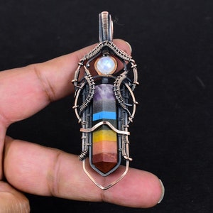 Op de afbeelding: Een hanger omwikkeld met koperdraad met een regenboog van zeven chakrastenen, waaronder amethist, lapis lazuli, citrien, carneol, rode jaspis en een maansteen bovenaan.